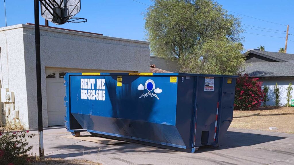 A blue dumpster with a painted message that reads, "rent me" with the roll off business phone number, dropped on a residential driveway