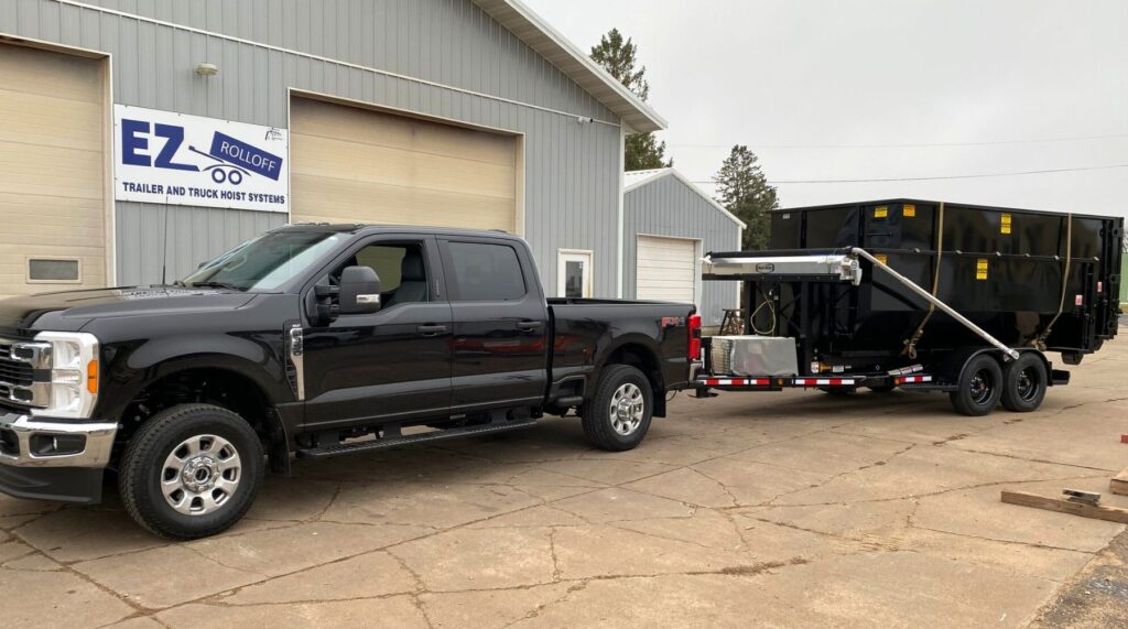 Black pickup truck hitched to a roll-off trailer; dumpsters secured with ratchet straps