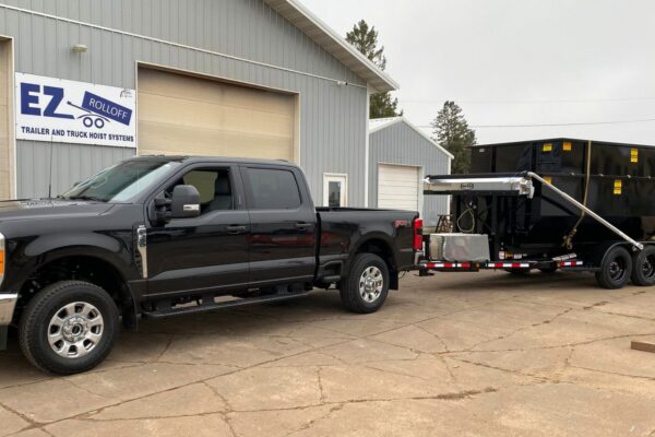 Black pickup truck hitched to a roll-off trailer; dumpsters secured with ratchet straps